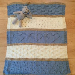 May include: A blue and cream knitted baby blanket with a brown stuffed bunny on top. The blanket has a variety of knit patterns, including hearts and stripes.