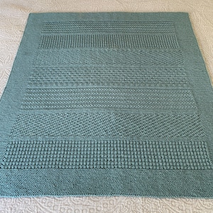 May include: A teal blue knitted blanket with a textured pattern. The blanket is made with a variety of stitches, creating a unique and interesting design.