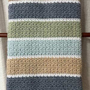 Op de afbeelding: Een gestreepte gehaakte babydeken in de kleuren beige, grijs, groen en blauw. De deken is gemaakt met een eenvoudige steek en heeft een witte rand.
