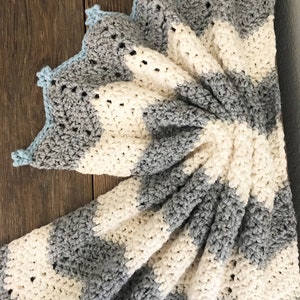 May include: A crocheted baby blanket with a striped pattern in white and gray. The blanket has a blue crocheted border.