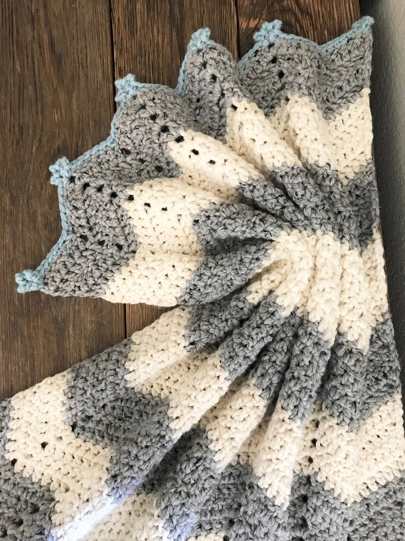 May include: A crocheted baby blanket with a striped pattern in white and gray. The blanket has a blue crocheted border.