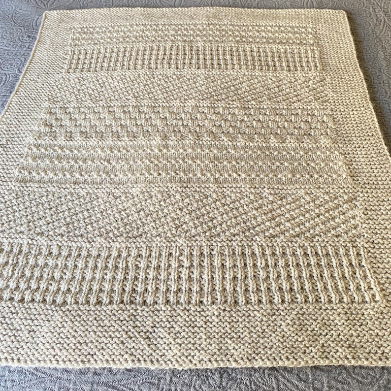May include: A beige knitted blanket with a textured pattern. The blanket is made with a variety of stitches, creating a unique and interesting design.