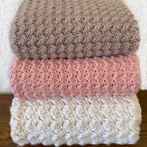 May include: Three crocheted blankets stacked on top of each other. The top blanket is brown, the middle blanket is pink, and the bottom blanket is white.