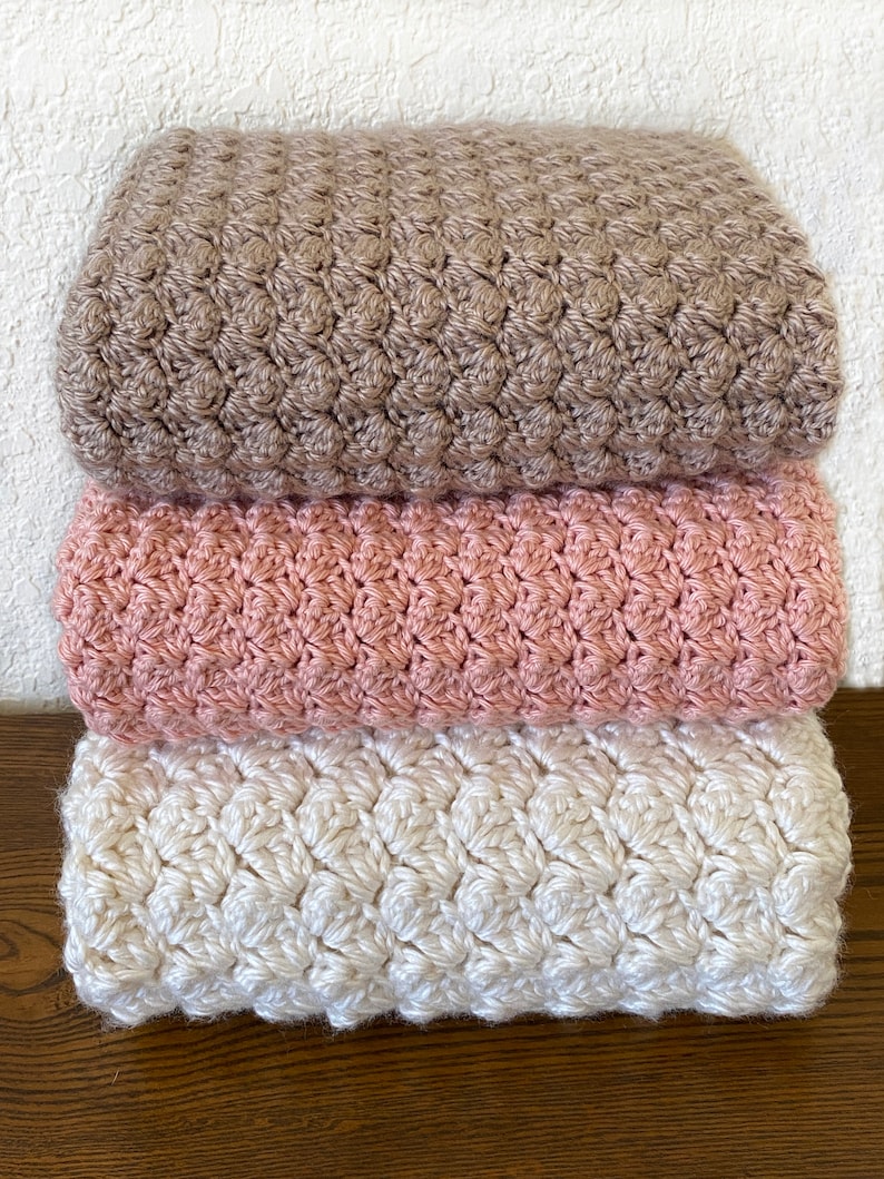 May include: Three crocheted blankets stacked on top of each other. The top blanket is brown, the middle blanket is pink, and the bottom blanket is white.