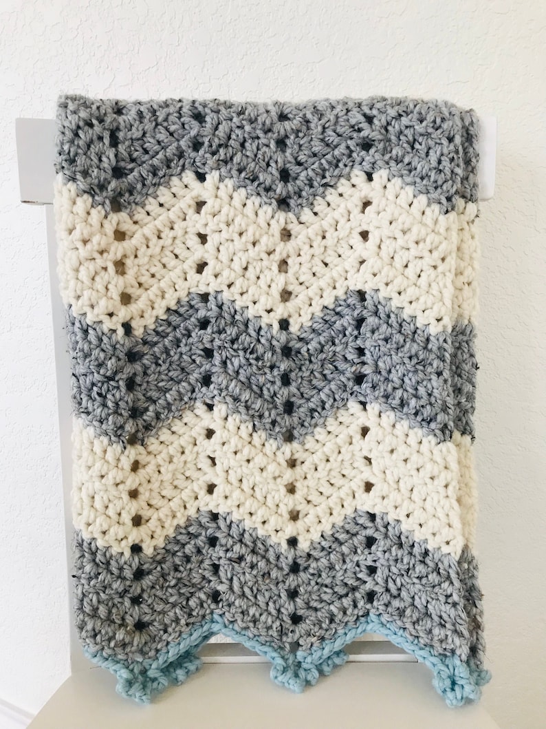 May include: A crocheted baby blanket with a chevron pattern in gray, white, and light blue. The blanket has a scalloped edge.
