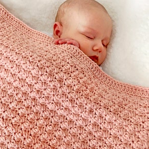 May include: A soft, pink crocheted baby blanket with a delicate stitch pattern. The blanket is draped over a sleeping baby.