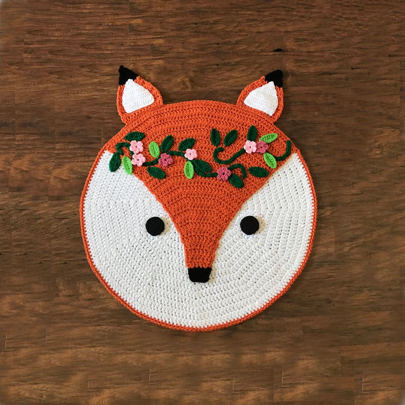 May include: A crocheted fox rug with a white face and orange fur. The fox is wearing a flower crown with pink and green flowers.