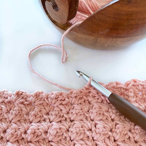 May include: A close-up of a pink crocheted blanket being worked on with a crochet hook and yarn. The yarn is coming from a wooden yarn bowl.