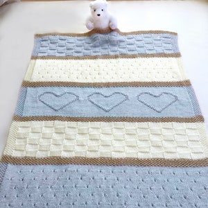 May include: A knitted baby blanket with a light blue, cream, and brown striped pattern. The blanket features a heart design and is made with a soft, textured yarn.