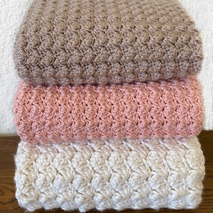 May include: Three crocheted blankets stacked on top of each other. The top blanket is brown, the middle blanket is pink, and the bottom blanket is white.