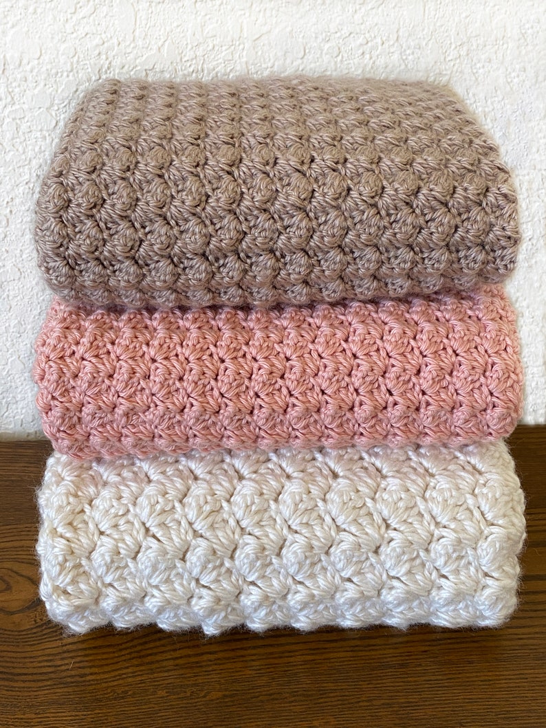May include: Three crocheted blankets stacked on top of each other. The top blanket is brown, the middle blanket is pink, and the bottom blanket is white.