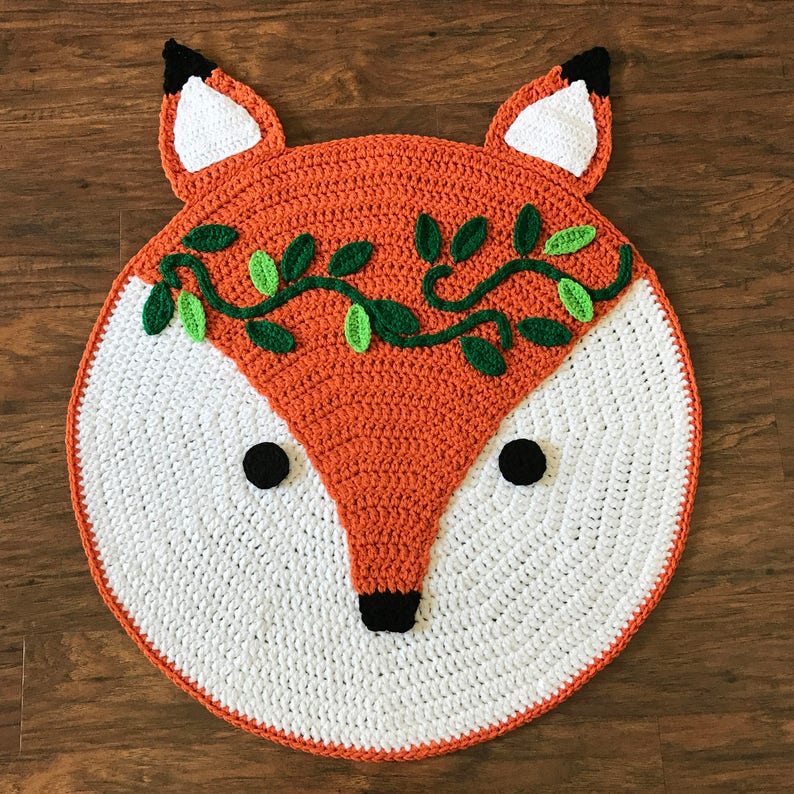 May include: A crocheted fox rug with a white background and orange fox face. The fox has green vines with leaves around its head.