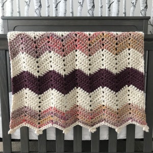May include: A white, purple, and pink chevron crochet baby blanket with a scalloped edge.
