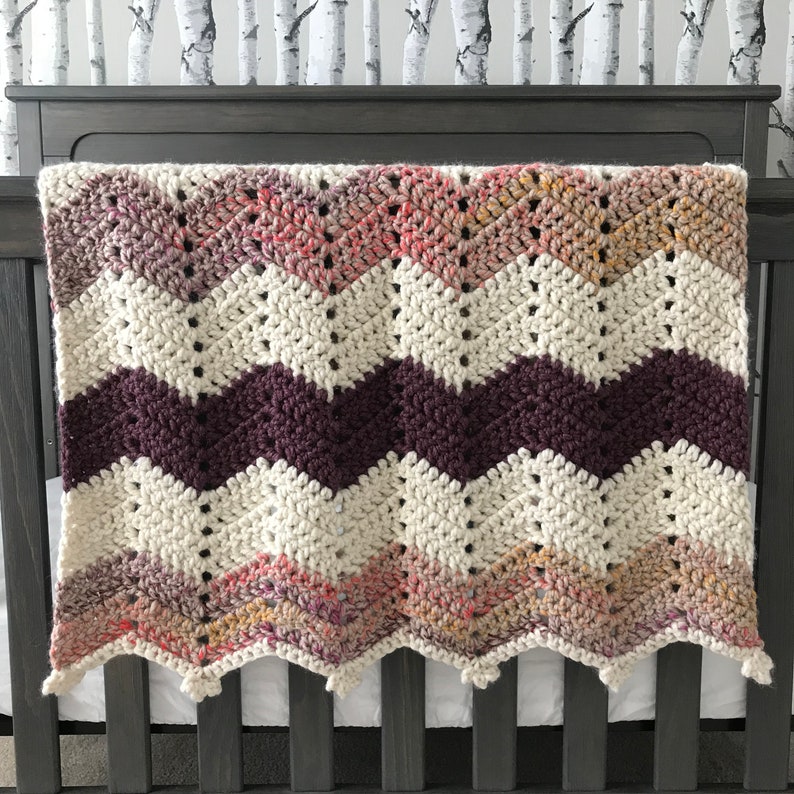 May include: A white, purple, and pink chevron crochet baby blanket with a scalloped edge.
