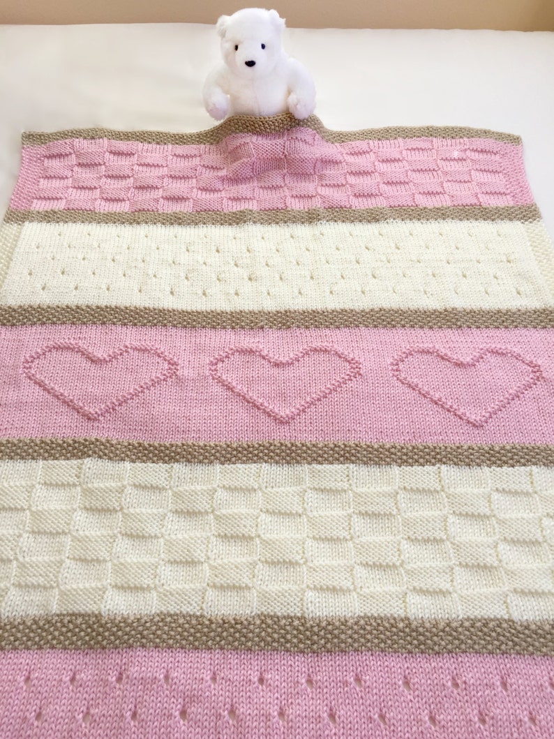 May include: A pink and white knitted baby blanket with a brown stripe and a white teddy bear peeking out from the top.