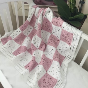 May include: A pink and white crocheted baby blanket with a square pattern. The blanket is made with a delicate, lacy stitch and has a scalloped edge.