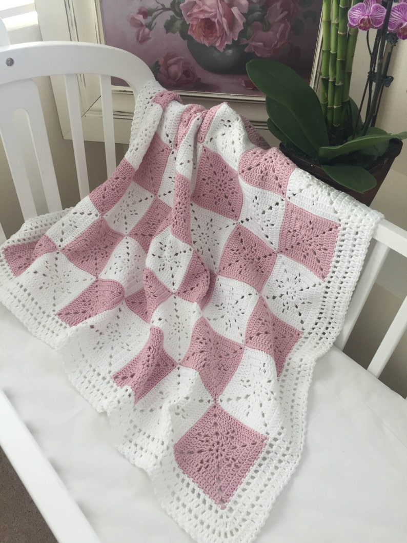 May include: A pink and white crocheted baby blanket with a square pattern. The blanket is made with a delicate, lacy stitch and has a scalloped edge.