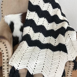 May include: Black and white chevron crochet throw blanket with black tassels. The blanket is draped over a chair.