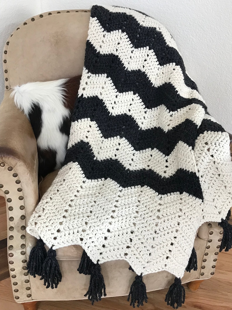 May include: Black and white chevron crochet throw blanket with black tassels. The blanket is draped over a chair.