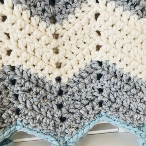 May include: A crocheted baby blanket with a chevron pattern in shades of gray, white, and light blue. The blanket has a scalloped edge.