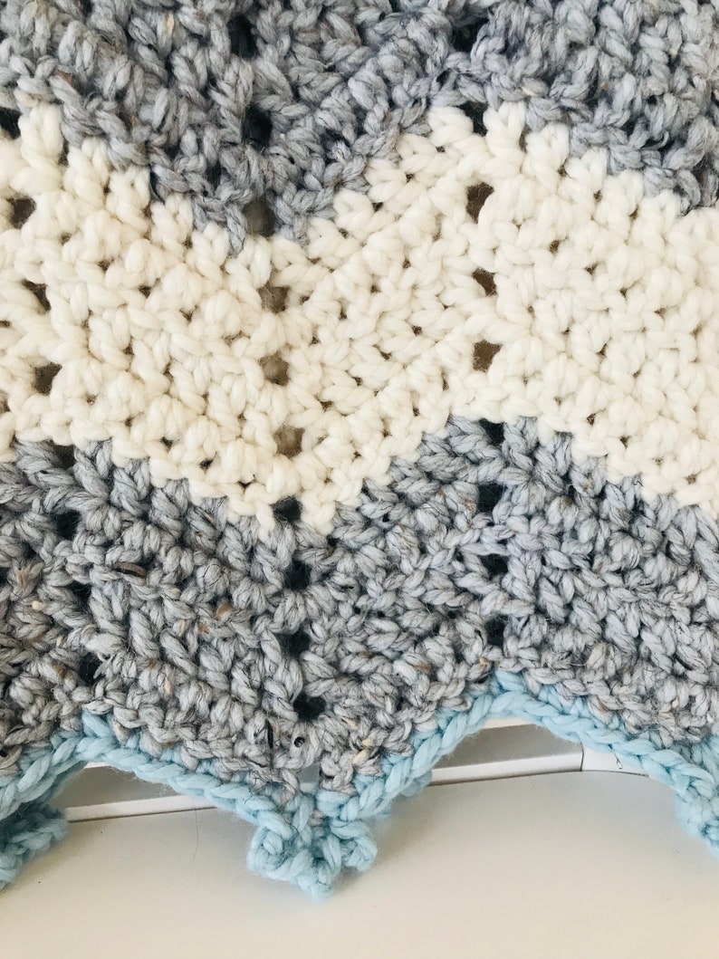 May include: A crocheted baby blanket with a chevron pattern in shades of gray, white, and light blue. The blanket has a scalloped edge.