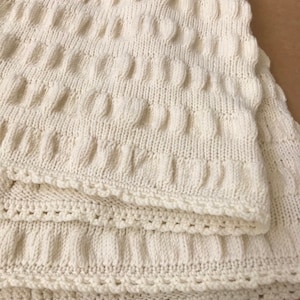 Könnte beinhalten: Cremefarbene Strickdecke mit einem strukturierten Muster aus erhabenen Kreisen und vertikalen Linien. Die Decke ist gefaltet und zeigt Schichten des weichen, warmen Materials. Die Kanten sind mit einem dekorativen, gewellten Rand versehen.