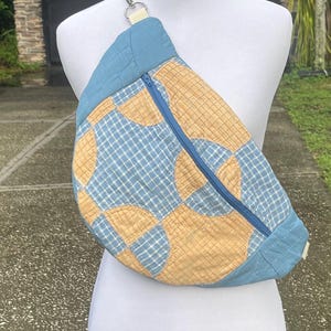 May include: A quilted sling bag featuring a blue and tan checkered pattern with circular motifs. The bag has a solid blue quilted top and bottom section, a diagonal blue zipper closure, and a cream-colored fabric strap attached with a silver metal clip.