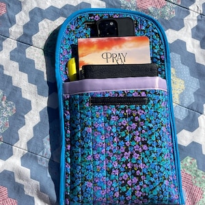 May include: A quilted fabric organizer with a floral pattern in shades of blue and purple. The organizer has a bright blue trim and contains a phone, a book with the word "PRAY", and other items. The background is a patterned quilt.