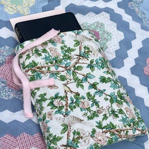 May include: A floral book sleeve with a pink ribbon closure. The white fabric features a pattern of birds, pink flowers, and green leaves. A black book and a pink book are inside the sleeve. The background is a blue and white quilt.