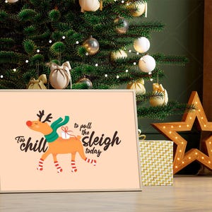 Puede incluir: Un cuadro enmarcado con un reno de dibujos animados con una bufanda verde y un regalo, con el texto "Too chill to pull the sleigh today". El cuadro se muestra frente a un árbol de Navidad y regalos.