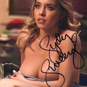 May include: A glossy photograph featuring a portrait of Sydney Sweeney with blonde hair, wearing a light-colored garment, and adorned with a delicate necklace featuring a small pendant. The image includes a prominent black ink autograph signature.
