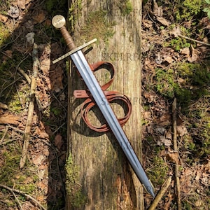 May include: A metal sword with a brown leather-wrapped grip and a circular pommel featuring a cross. The crossguard is engraved with the Latin text IN HOC SIGNO VINCES. A brown leather strap is coiled behind the blade, which has a central fuller.