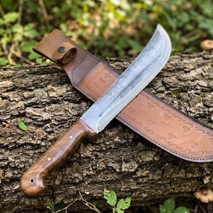 May include: A fixed-blade knife with a curved steel blade and a polished brown wooden handle featuring two brass rivets. The knife is accompanied by a tan leather sheath that displays an embossed decorative pattern and a snap button closure.