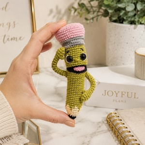May include: A crocheted pencil plush toy featuring a green body, a pink eraser top with a grey band, and a tan tip. The character has black safety eyes, a wide open mouth with a pink tongue, and two arms, one of which is raised in a salute.