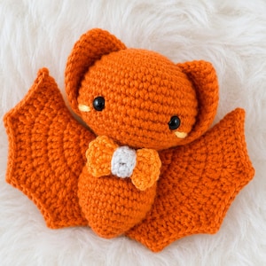 Puede incluir: Un juguete de murciélago tejido a ganchillo en un color naranja vibrante. El murciélago presenta orejas grandes y redondeadas, dos pequeños ojos de plástico negro con detalles amarillos bordados debajo y una pajarita naranja y blanca.