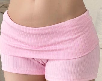 May include: A pair of light pink ribbed shorts featuring a low-rise waistband with a folded-over design. The garment is constructed from a soft, textured fabric with vertical ribbing throughout, providing a snug fit around the hips and upper thighs.