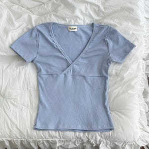 May include: A light blue short-sleeve top made of ribbed fabric featuring a V-neckline with a wrap-style front detail. The garment includes a horizontal seam across the midsection and a small white rectangular label at the neck with black text.