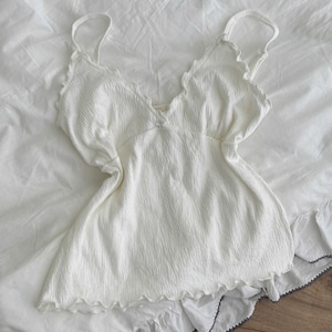 Könnte beinhalten: Ein ärmelloses weißes Camisole-Top aus strukturiertem, geknittertem Stoff. Das Kleidungsstück hat einen tiefen V-Ausschnitt mit zarten Rüschenkanten, dünne Spaghettiträger und einen leicht ausgestellten Saum, der ebenfalls einen dezenten Rüschenbesatz aufweist.