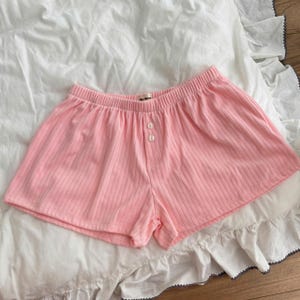 May include: Light pink ribbed shorts featuring an elasticized waistband and a front placket with two white buttons. The garment is made of a soft, textured fabric and includes a small interior brand label at the center of the waistband.