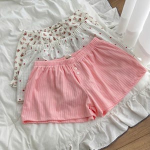 May include: Three pairs of lounge shorts with elastic waistbands and two front buttons. The front pair is solid light pink with a ribbed texture. The middle pair features a white grid pattern with small red dots. The back pair has a floral print.