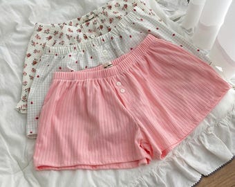 May include: Three pairs of lounge shorts with elastic waistbands and two front buttons. The front pair is solid light pink with a ribbed texture. The middle pair features a white grid pattern with small red dots. The back pair has a floral print.