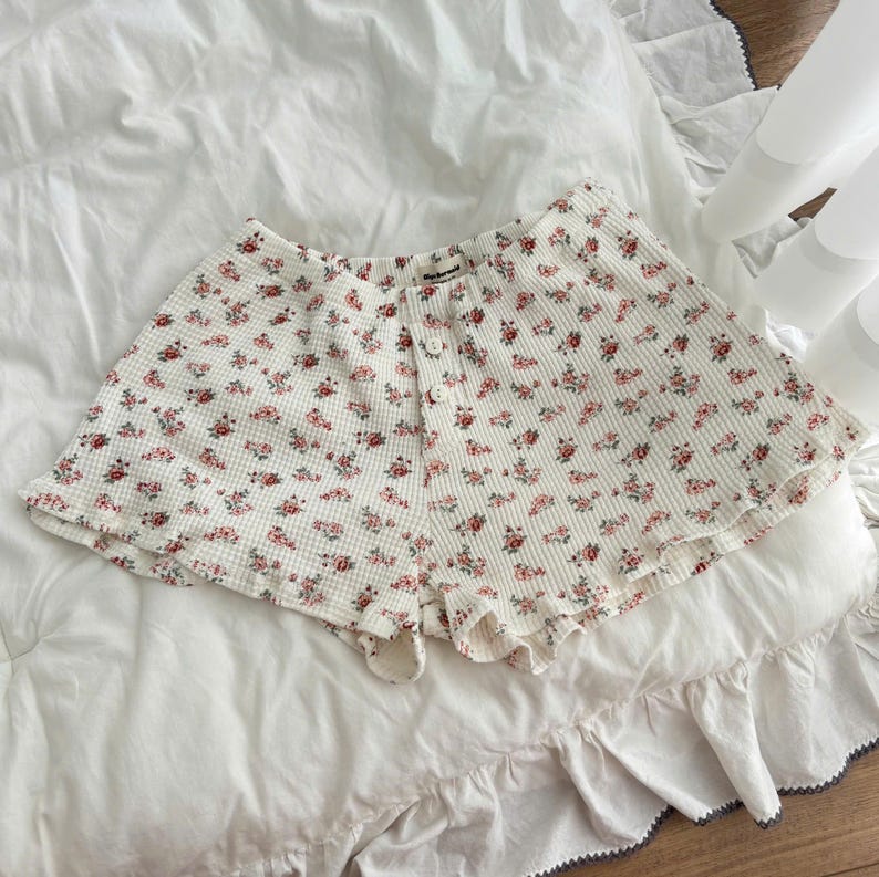May include: Off-white waffle knit shorts featuring a small red and green floral pattern. The shorts have a high-waisted cut with a front button placket containing three white buttons and a small fabric label at the center of the waistband.