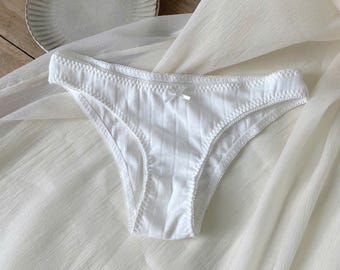 May include: A pair of white underwear featuring a subtle vertical ribbed texture, a scalloped lace trim along the waistband and leg openings, and a small centered bow detail at the front. The garment is crafted from a soft, lightweight fabric.