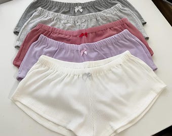 May include: A stack of five pairs of ribbed shorts in white, lavender, dusty rose, light gray, and dark gray. Each pair features an elastic waistband and a small decorative bow centered at the front. The shorts have a soft, textured fabric finish.