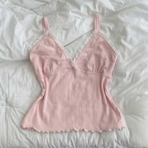 Puede incluir: Una camiseta de tirantes de color rosa claro con textura acanalada y un escote en V profundo adornado con delicado encaje blanco. La prenda tiene tirantes finos y un dobladillo con volantes, confeccionada en tela suave con canalé vertical.