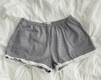 May include: Grey lounge shorts featuring a soft, heathered fabric texture, an elasticized waistband with a small interior tag, and delicate white lace trim detailing along the curved leg openings. The shorts have a relaxed fit and simple construction.