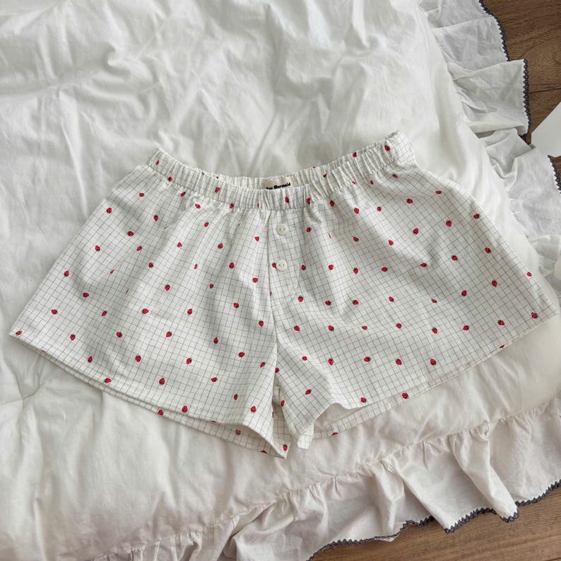 May include: White shorts featuring a thin black grid pattern and a repeating small red strawberry print. The shorts have an elastic waistband and a front placket with two white buttons. The fabric is lightweight and the cut is loose and relaxed.