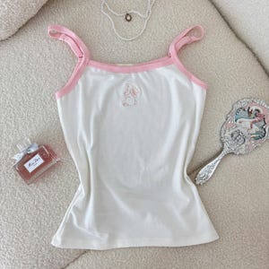 May include: A white sleeveless vest top with pink trim along the neckline and straps, featuring a small embroidered pink rabbit outline in the centre. The garment is displayed alongside a pearl necklace, a perfume bottle, and a hand mirror.