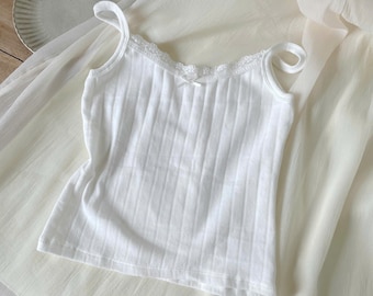 May include: A white ribbed camisole featuring thin shoulder straps, a scoop neckline trimmed with delicate white lace, and a small center bow detail. The garment is crafted from a textured ribbed fabric, providing a fitted silhouette.