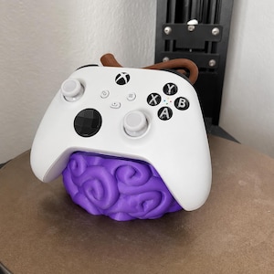 May include: A white Xbox wireless controller resting on a purple, brain-shaped stand with a brown, stem-like protrusion at the top. The controller features standard black buttons, white thumbsticks, and a black directional pad, all mounted on the base.